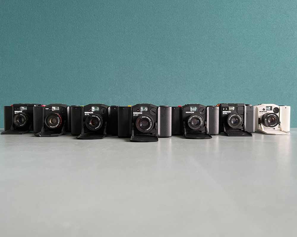Sieben Minox 35 Kameras in einer Reihe vor einer blauen Wand.